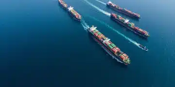 An aerial drone view emphasizing global supply chain disruption, with a container ship subtly blocking a shipping lane, causing a geometric pattern of other ships and a visible ripple effect across the serene ocean.