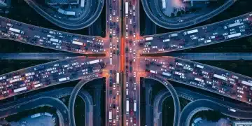 Aerial view of a sprawling highway interchange at dusk, choked with traffic forming geometric patterns, with a central section closed by orange barriers, symbolizing widespread disruption from a global event.